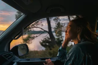 A woman sits on the passenger seat of a moving camper during winter in Portugal. You see the sunset outside.